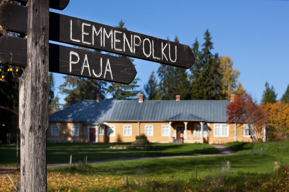 Musta puinen opasteviitta, jossa on valkoisella tekstit "LEMMENPOLKU" ja "PAJA". Taustalla näkyy epätarkkana Möhkön ruukkimuseon keltainen Pytinki-rakennus ja aurinkoinen nurmipiha.