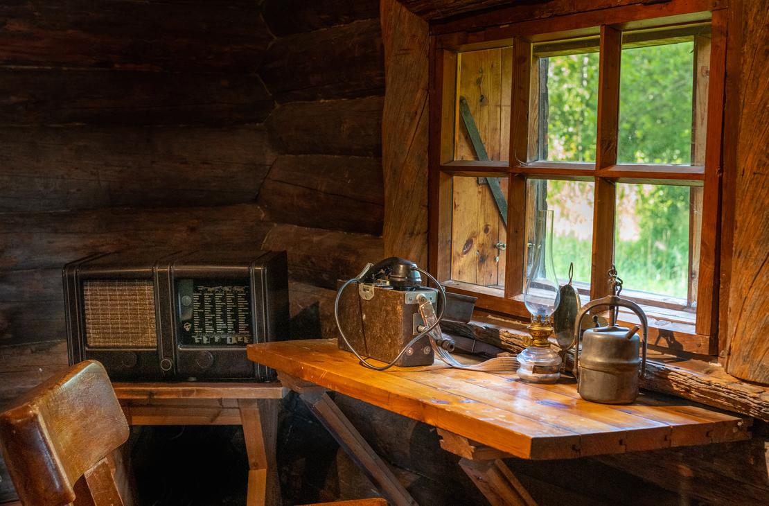 Kuva Rajakenraalin majan sisätilasta, jossa esitetään perinteistä hirsistä sisustusta. Lähikuva keskittyy puiseen ikkunanpöytään, jolla on vanhanaikainen termospullo ja metallinen astia. Pöytä on sijoitettu ikkunan eteen, josta siivilöityy sisään valoa ja näkyy ulkoa vihreää kasvillisuutta. Vasemmalla tumman hirsiseinän vieressä näkyy vanha pöytäradio.