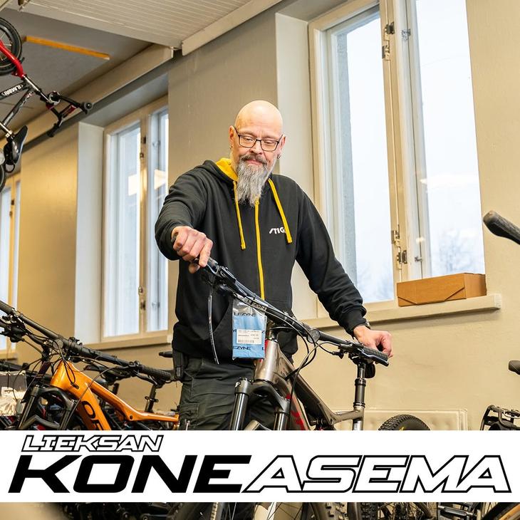 Silmälasipäinen, parrakas mies tarkastelemassa polkupyörää myymälässä. Kuvan alalaidassa on Lieksan Koneasema -tekstilogo.