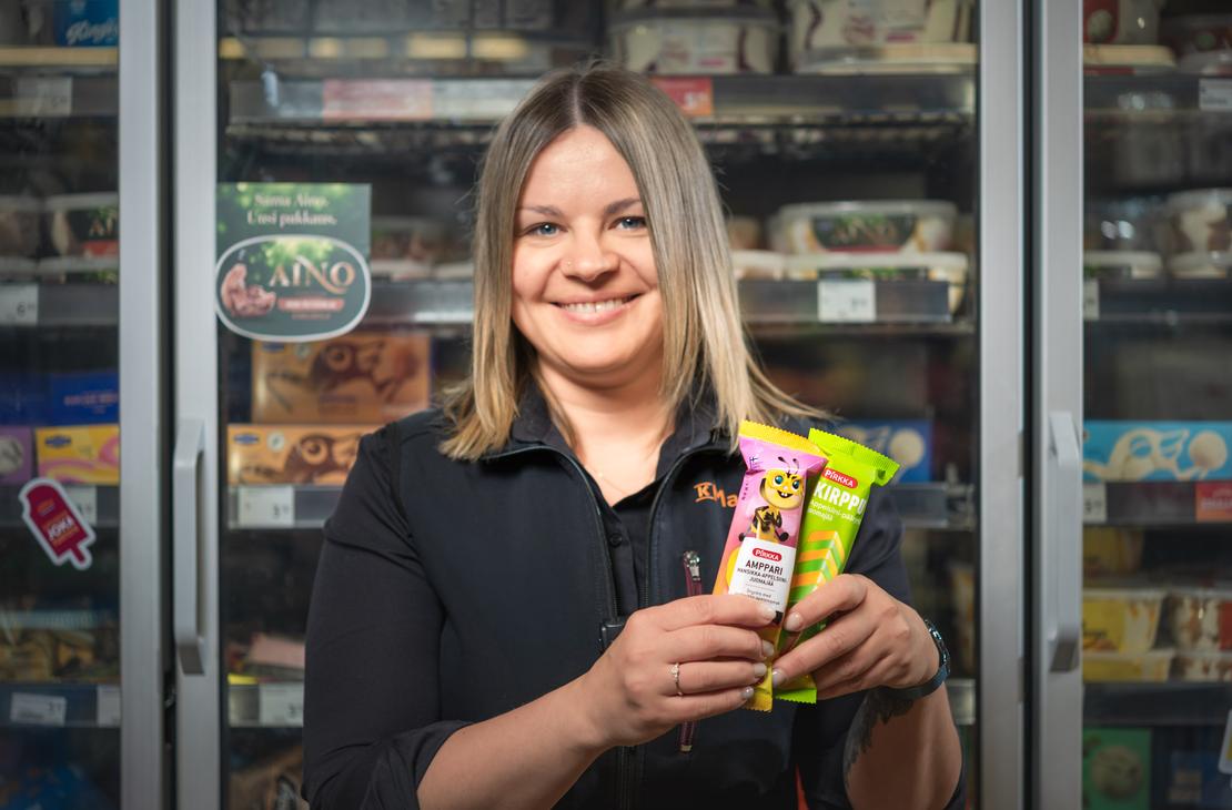 Mustaan K-Market-paitaan pukeutunut ty&ouml;ntekij&auml; hymyilee ja pitelee k&auml;siss&auml;&auml;n Pirkka Amppari- ja Pirkka Kirppu -mehuj&auml;it&auml; K-Market Ilomantsissa. H&auml;nen takanaan on lasiovinen pakastekaappi, jonka hyllyill&auml; on erilaisia j&auml;&auml;tel&ouml;it&auml; ja jonka ovessa on py&ouml;re&auml; Aino-j&auml;&auml;tel&ouml;n tarra.