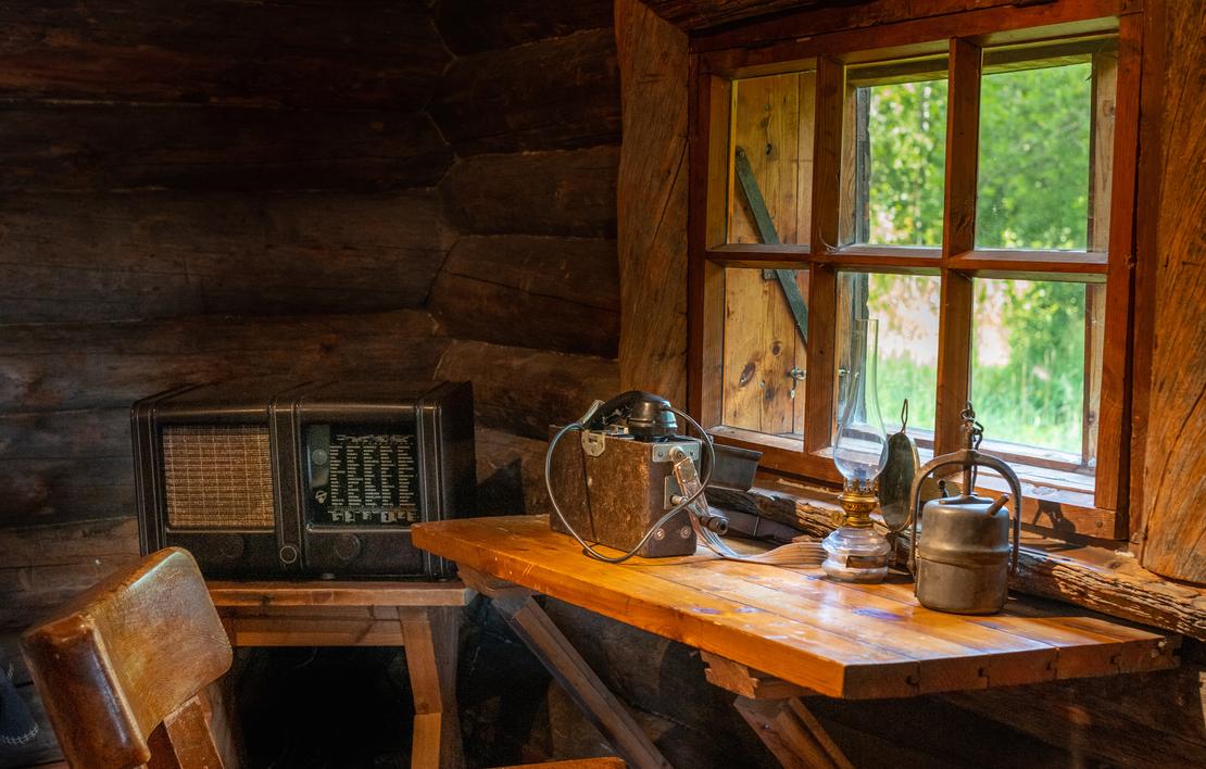 Kuva Rajakenraalin majan sisätilasta, jossa esitetään perinteistä hirsistä sisustusta. Lähikuva keskittyy puiseen ikkunanpöytään, jolla on vanhanaikainen termospullo ja metallinen astia. Pöytä on sijoitettu ikkunan eteen, josta siivilöityy sisään valoa ja näkyy ulkoa vihreää kasvillisuutta. Vasemmalla tumman hirsiseinän vieressä näkyy vanha pöytäradio.