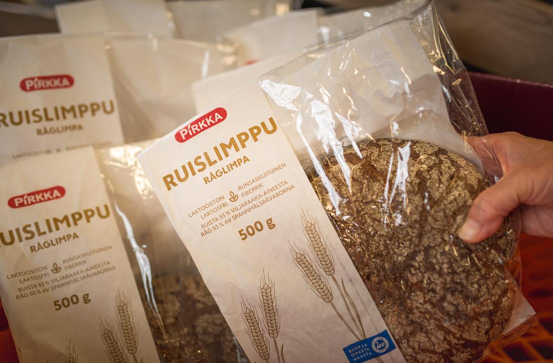 Käsi pitelee osittain läpinäkyvää Pirkka Ruislimppu -leipäpakkausta K-Market Ilomantsissa. 500 gramman pakkauksesta erottuu pyöreä ruisleipä sekä tekstit ”Pirkka Ruislimppu Råglimpa”. Taustalla on useita muita samanlaisia leipäpakkauksia rivissä.