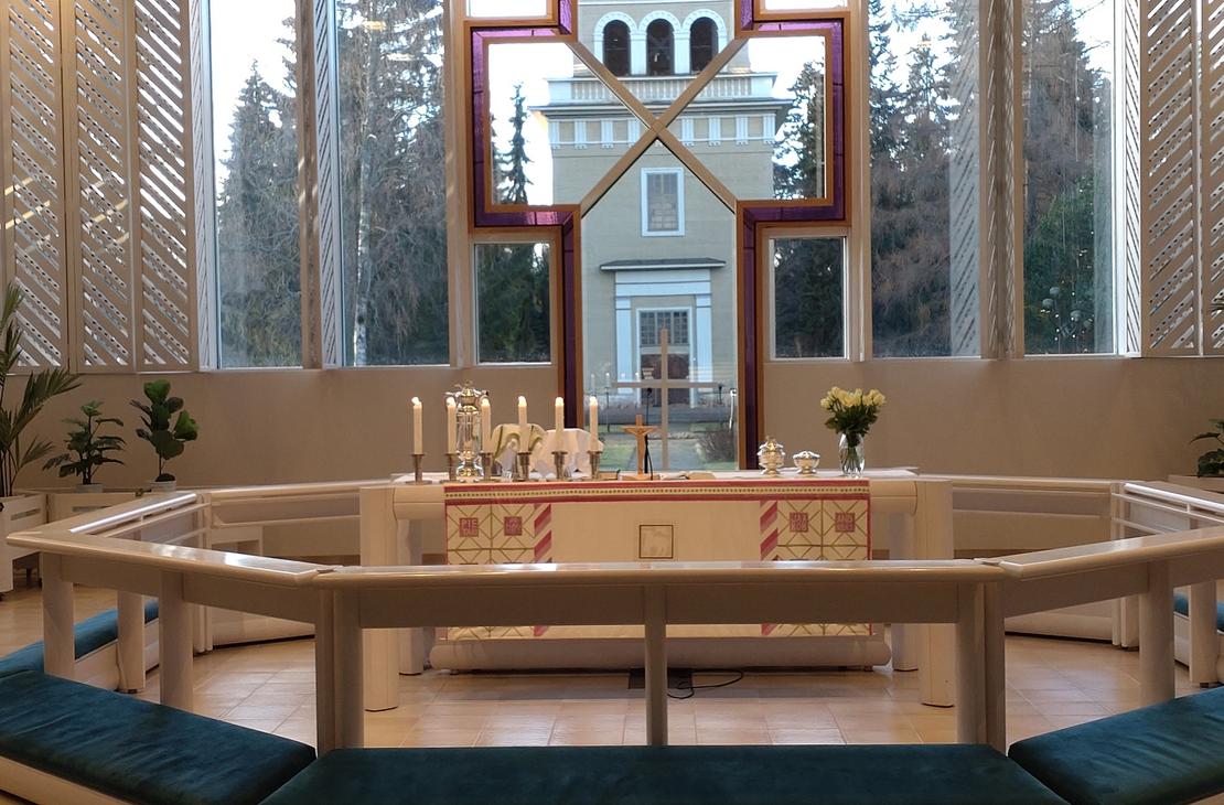 Lieksan kirkon moderni alttari, jonka takana olevasta suuresta ristikuvioisesta ikkunasta avautuu näkymä vanhaan kellotapuliin. Alttarilla on kynttilöitä ja kukkia.