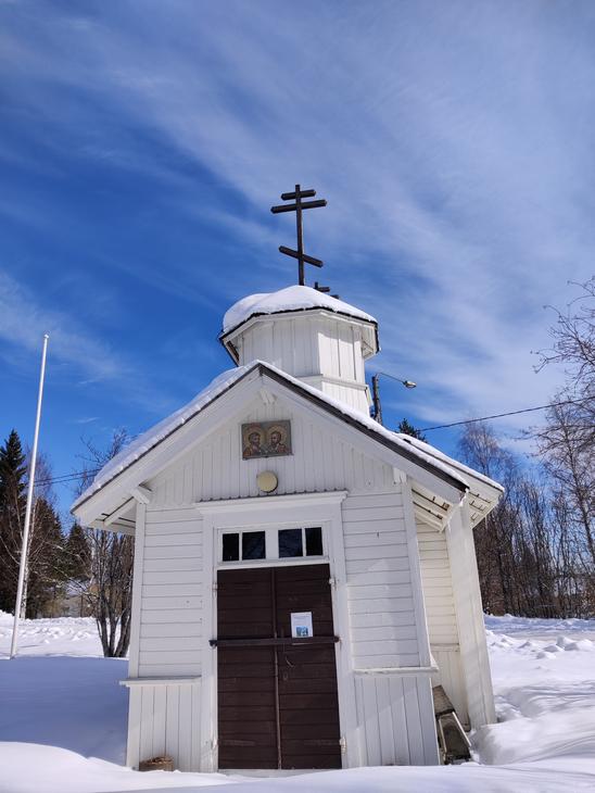 Pieni valkoinen puurakennus, mahdollisesti tsasouna, jonka katolla on ortodoksinen risti. Rakennus sijaitsee lumisessa ympäristössä kirkkaan sinisen taivaan alla.