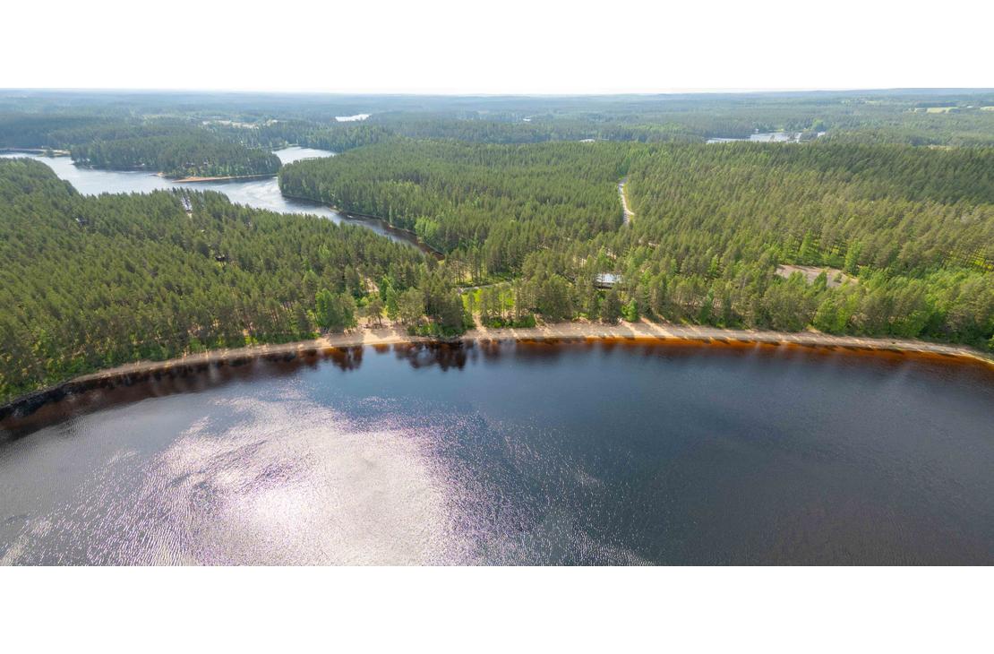 Korkealta otettu laaja panoraamailmakuva Suomen järvi- ja metsämaisemasta. Etualalla on kaareva hiekkaranta ja sen takana silmänkantamattomiin jatkuvaa tiheää havumetsää ja vesistöjä.