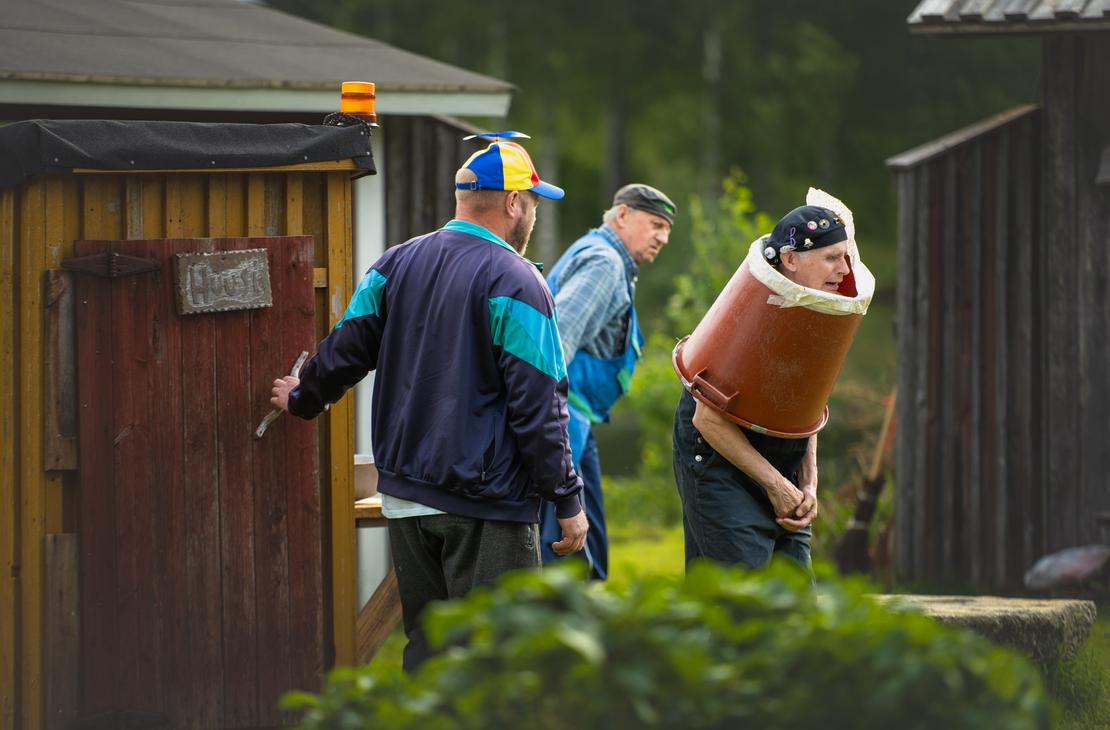 Möhkön kesäteatterin Peitetarina-näytelmän kohtaus, jossa kolme miestä esiintyy ulkonäyttämöllä. Vasemmalla mies, jolla on värikäs potkurilippis, on avaamassa puisen ulkokäymälän ovea, jossa on teksti "Huussi". Oikealla vanhempi mies seisoo vartalonsa ympärille asetetun suuren ruskean muovisaavin sisällä ilmehtien vaivautuneesti, ja heidän takanaan kolmas mies seuraa tilannetta.