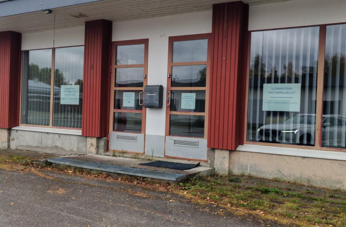 Ilomantsin Yrityspalvelut -toimiston sisäänkäynti ulkoapäin. Yksikerroksisen rakennuksen julkisivussa on valkoista seinää, pystysuuntaisia punaruskeita puupaneeleita sekä suuria ikkunoita pystylamellikaihtimilla. Keskellä on kaksi ruskeakehyksistä lasiovea, joiden välissä on harmaa postilaatikko ja edessä metalliritiläinen askelma mattoineen. Oikeanpuoleisessa ikkunassa on valkopohjainen kyltti, jossa lukee "ILOMANTSIN YRITYSPALVELUT KIRJANPITO - PALKANLASKENTA - VEROASIAT". Etualalla on asfaltoitu piha-alue, ja rakennuksen seinustalla kasvaa hieman ruohoa.