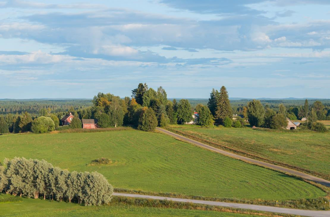 Ilmakuva kumpuilevasta peltomaisemasta ja sen läpi kulkevasta maantiestä. Rinteessä on punaisia ja valkoisia maalaistaloja sekä puurivistöjä.