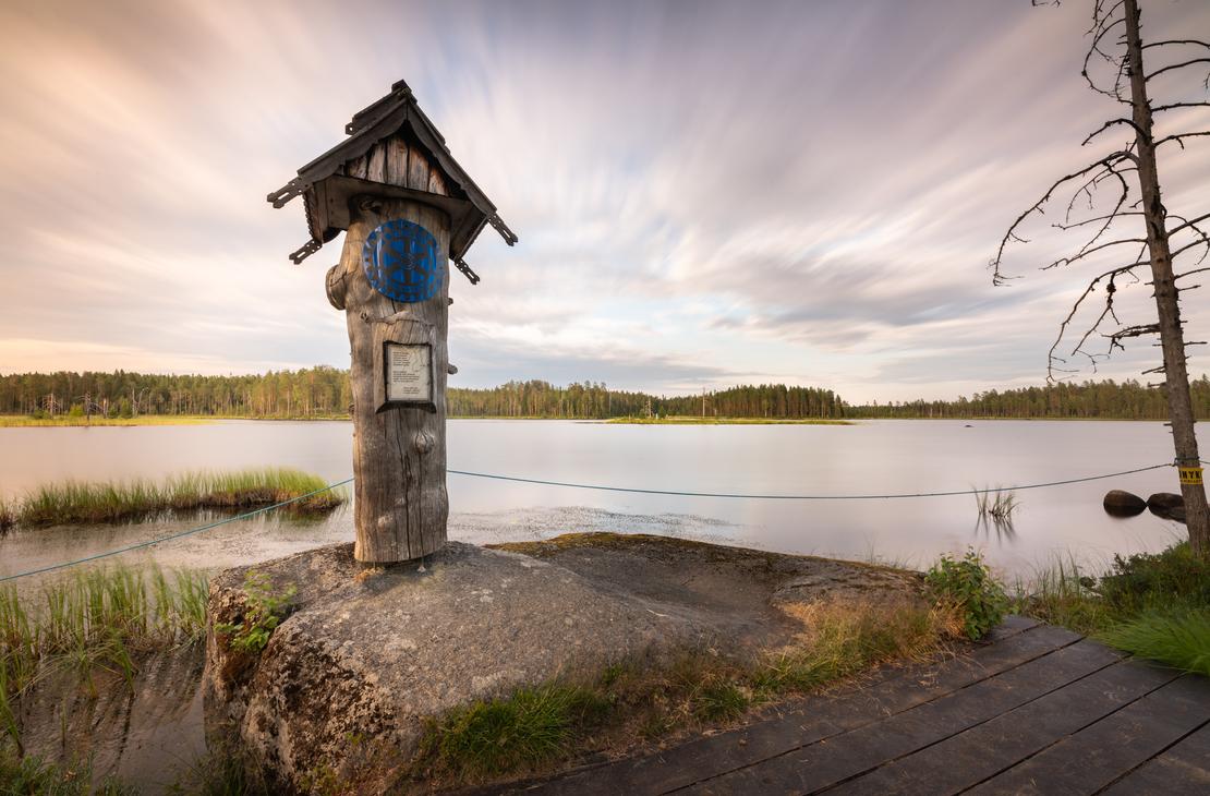 Kuva Suomen it&auml;isimm&auml;lt&auml; pisteelt&auml; auringonlaskun tai -nousun aikaan. Etualalla on puinen, veistetty muistomerkki tai pylv&auml;s, jonka p&auml;&auml;ll&auml; on pieni katos ja sininen, py&ouml;re&auml; symboli. Muistomerkki seisoo kivetyll&auml; alustalla veden &auml;&auml;rell&auml;. Taustalla avautuu laaja j&auml;rvimaisema, jonka vastarannalla on tummaa mets&auml;&auml;. Taivas on dramaattinen, pilvinen, ja siin&auml; n&auml;kyy pitk&auml;n valotusajan aiheuttamaa liikett&auml;. Oikealla n&auml;kyy kuollut, karu puu ja osittain puinen k&auml;velysilta.