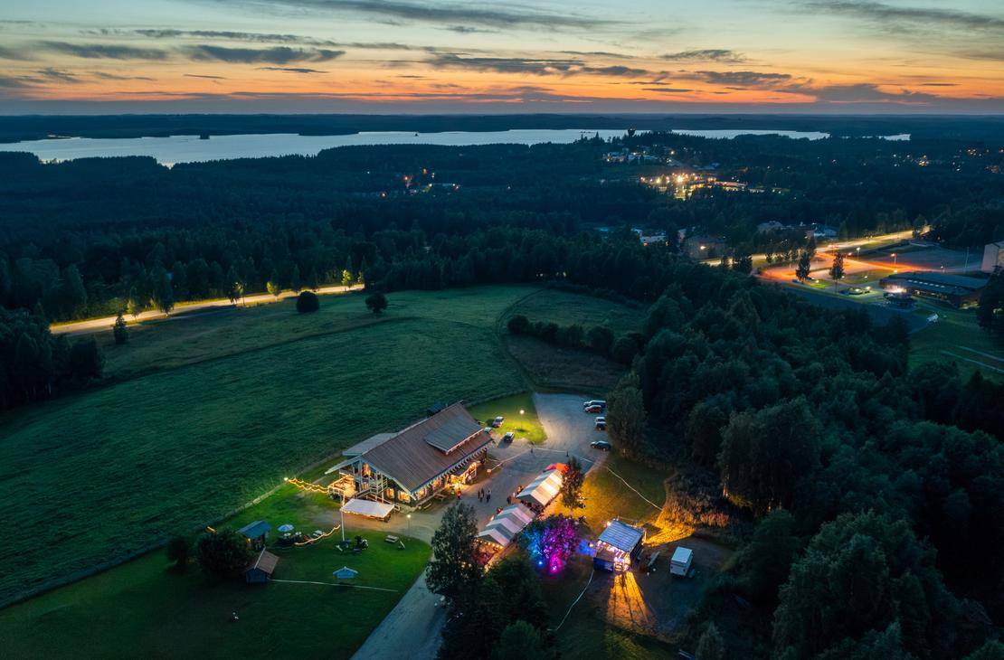Ilmakuva Parppeinpirtin valaistusta ravintolakokonaisuudesta iltahämärässä. Tummansininen metsämaisema ympäröi aluetta, ja taustalla siintää järvi ja asutuksen valoja auringonlaskun kajossa.