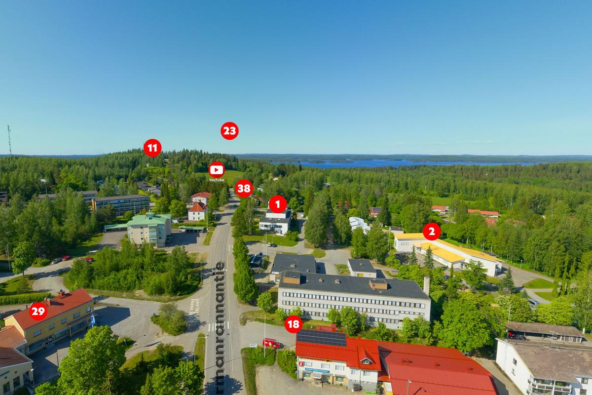 Näkymä 360 Palvelukartasta, joka esittää ilmakuvaa taajamasta aurinkoisena päivänä. Keskellä kulkee tie, jonka varrelle on merkitty useita kohteita punaisilla numerokuvakkeilla (kuten 1, 2, 11, 23) ja YouTube-logolla. Taustalla siintää järvi ja metsäisiä mäkiä.