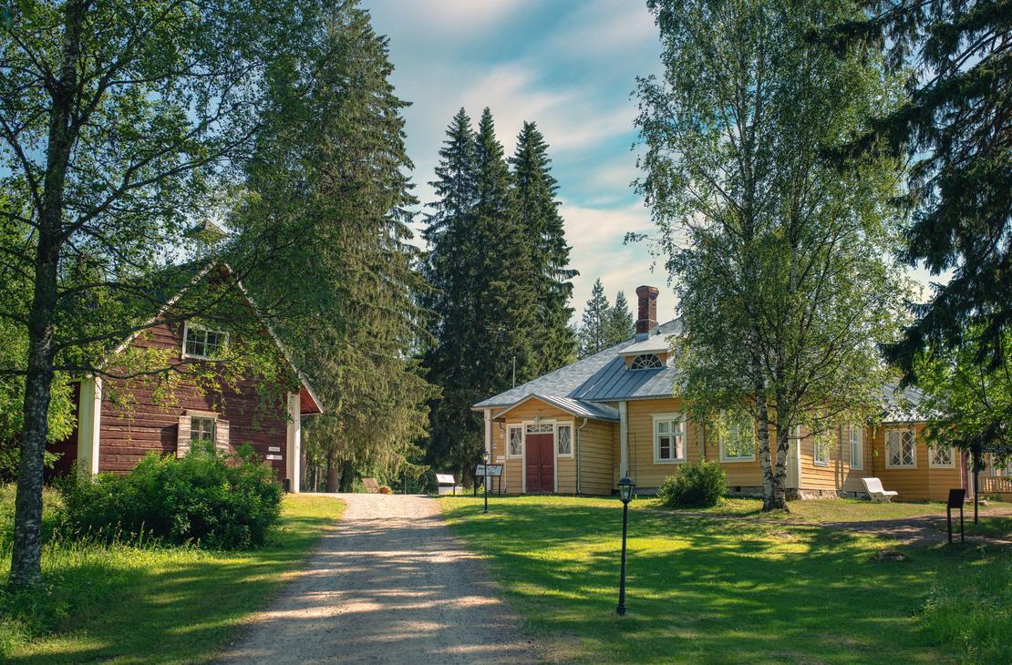 Kuva Möhkön ruukkimuseon alueelta esittää historiallista pihapiiriä. Oikealla on pitkä, matala, keltainen puutalo punaisella ovella ja harmaalla katolla. Vasemmalla näkyy korkeampi, punamultainen ulkorakennus tai aitta. Rakennusten välissä kulkee hiekkainen tie, ja niitä ympäröi vanhoja koivuja ja havupuita aurinkoisessa kesäympäristössä.