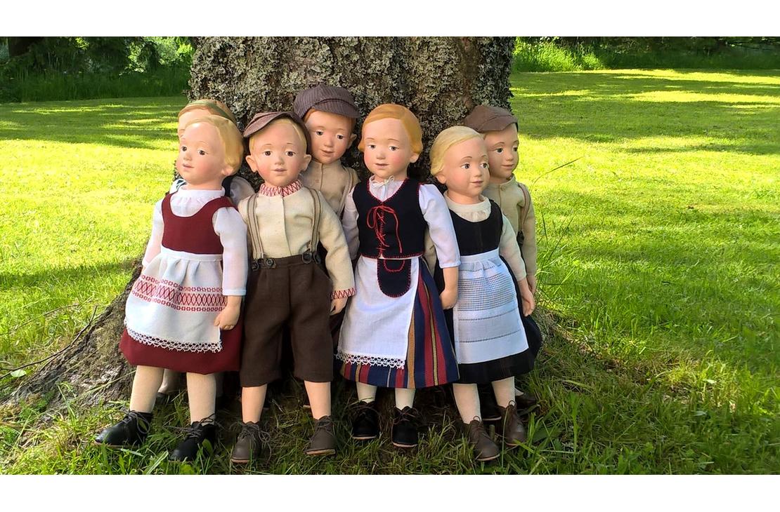 Kuva esittää kahta pehmolelua: vasemmalla on suuri, vaaleanruskea, perinteisen näköinen nallekarhu, jolla on yllään valkoinen paita ja siniset housut. Oikealla on pienempi nukke, jolla on punaiset hiukset ja yllään raidallinen paita ja tumma hame. Molemmat lelut istuvat pöydällä tai hyllyllä, ja tausta on epätarkka. Kuva on todennäköisesti peräisin Nukkemuseosta tai leluliikkeestä.