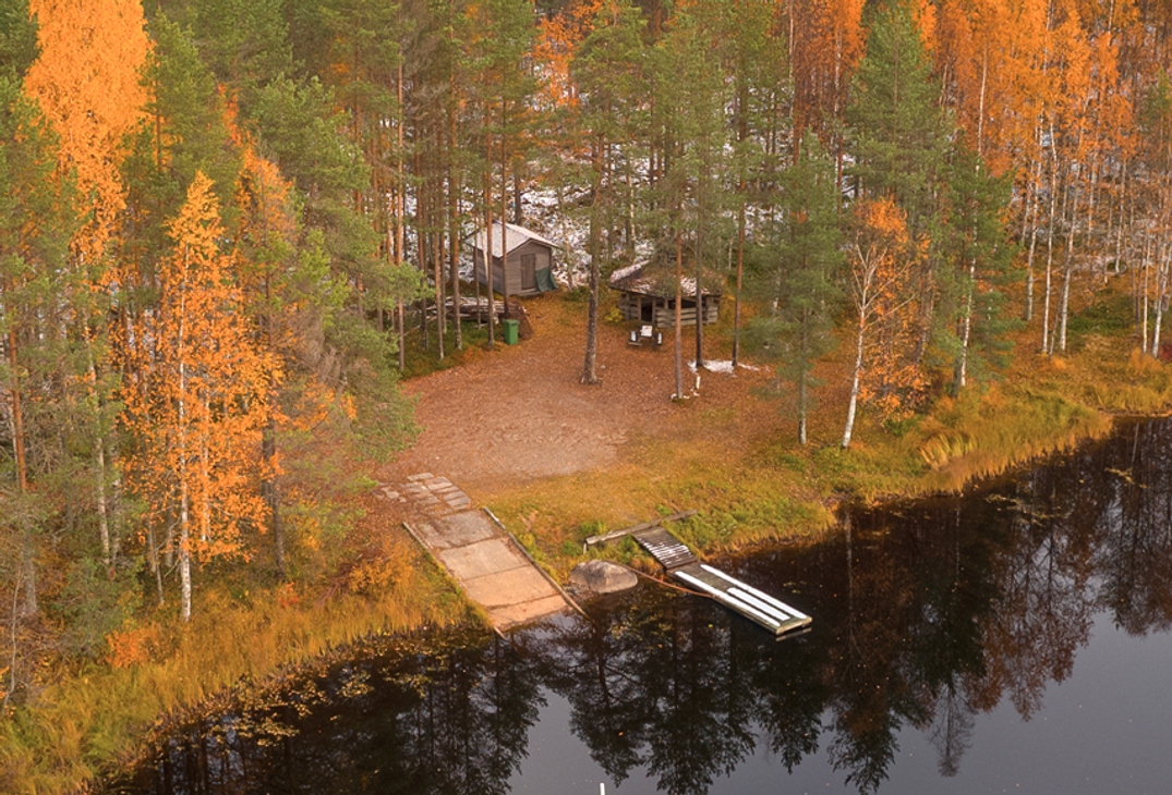 Ilmakuva tai korkealta otettu kuva syksyisest&auml; j&auml;rvimaisemasta, jossa rannalla on pieni m&ouml;kki tai leiripaikka. Ranta-alue on peitetty tummalla maalla/hiekalla ja sit&auml; ymp&auml;r&ouml;i mets&auml;, jonka puissa on voimakkaat keltaiset ja oranssit ruskan v&auml;rit. Kuvassa n&auml;kyy kaksi erilaista, puista laituria ulottumassa tummaan veteen: toinen leve&auml;mpi ja lyhyempi, toinen kapeampi ja pidempi.