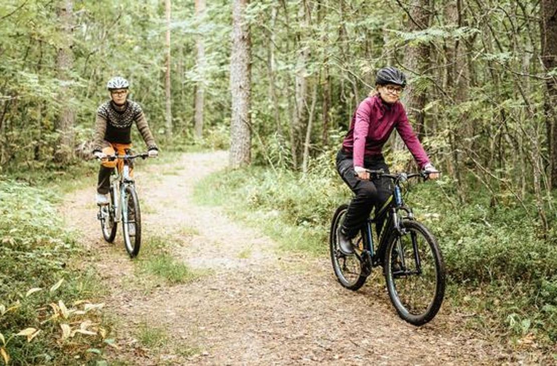 Kaksi henkilöä, nainen etualalla ja toinen henkilö taustalla, pyöräilevät mutkittelevaa hiekkatietä pitkin tiheässä metsässä. Nainen etualalla on pukeutunut pinkkiin paitaan ja kypärään, ja hän ajaa tummaa maastopyörää. Taustalla oleva pyöräilijä on pukeutunut tummiin vaatteisiin ja ajaa vaaleampaa pyörää. Polkua reunustaa runsas, vihreä aluskasvillisuus.