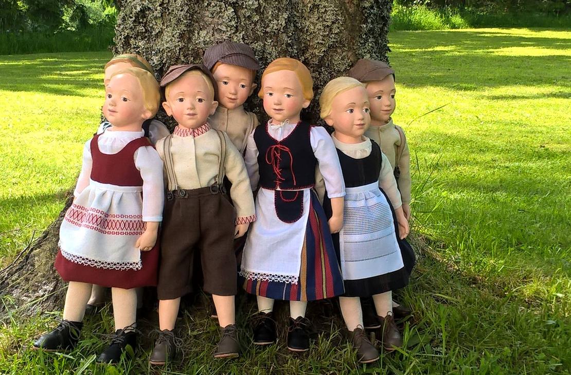 Kuva esittää kahta pehmolelua: vasemmalla on suuri, vaaleanruskea, perinteisen näköinen nallekarhu, jolla on yllään valkoinen paita ja siniset housut. Oikealla on pienempi nukke, jolla on punaiset hiukset ja yllään raidallinen paita ja tumma hame. Molemmat lelut istuvat pöydällä tai hyllyllä, ja tausta on epätarkka. Kuva on todennäköisesti peräisin Nukkemuseosta tai leluliikkeestä.