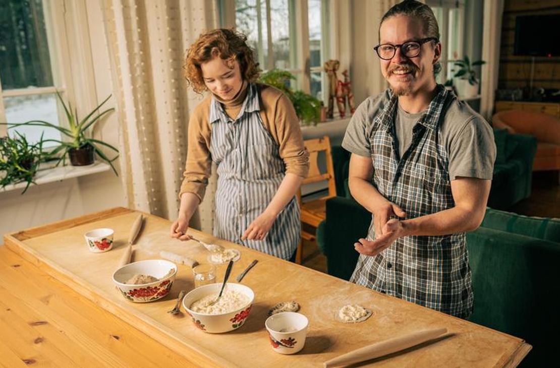 Sisäkuva, jossa kaksi aikuista, nainen ja mies, leipovat perinteisiä karjalanpiirakoita puisen pöydän ääressä. Molemmat henkilöt ovat pukeutuneet essuihin. Mies oikealla katsoo suoraan kameraan ja hymyilee. Pöydällä on taikinakulhoja ja valmiita, ohuita taikinapohjia. Tila on lämminhenkinen, ja taustalla näkyy ikkuna ja kasveja. Toiminta liittyy todennäköisesti Äksyt Ämmät -yrityksen karjalanpiirakka-työpajoihin tai -palveluihin.