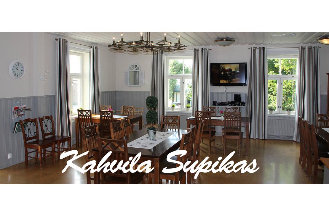 Kuva esittää Kahvila Supikkaan sisätiloja. Tila on valoisa ja siinä on useita suuria ikkunoita harmailla verhoilla. Seinät ovat kaksiväriset: alaosa on harmaa ja yläosa valkoinen. Huoneessa on useita puisia pöytiä ja tuoleja, ja pöydillä on valkoiset liinat. Oikealla on seinällä taulutelevisio. Kuvan alaosassa on suuri, valkoisella, kursivoidulla fontilla teksti "Kahvila Supikas".