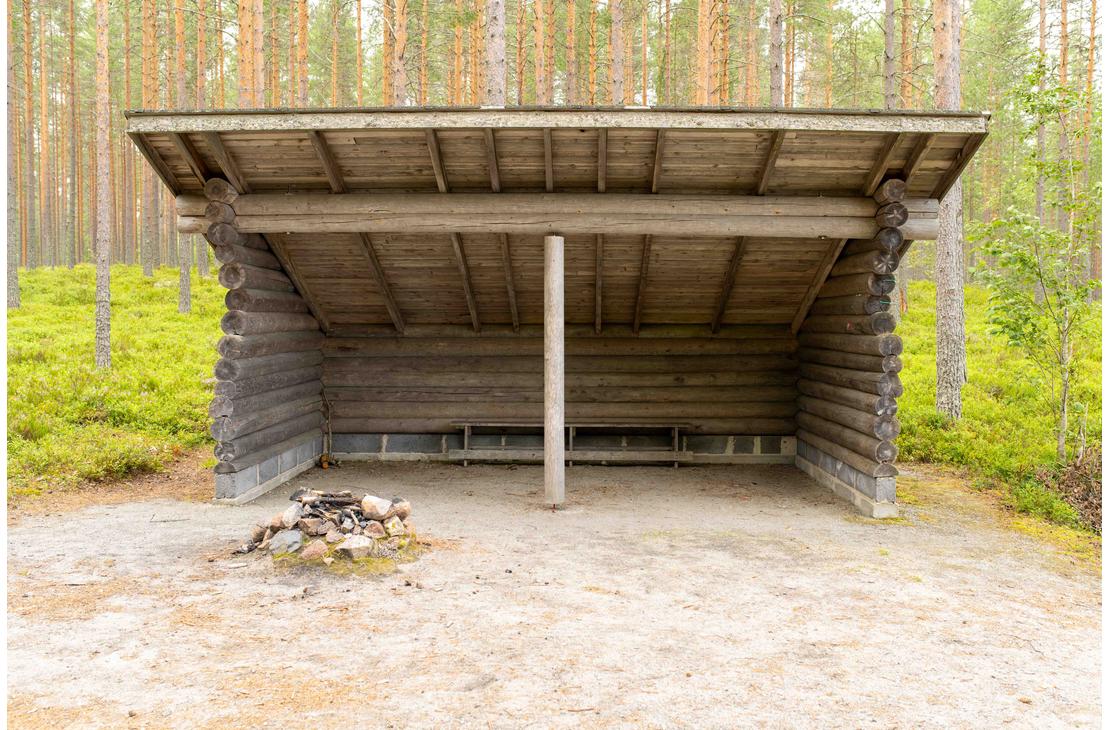 Suuri hirsirakenteinen laavu mäntymetsän siimeksessä. Laavun edustalla hiekalla on kivistä koottu nuotiopaikka ja sisällä pystytuki sekä puupenkki.