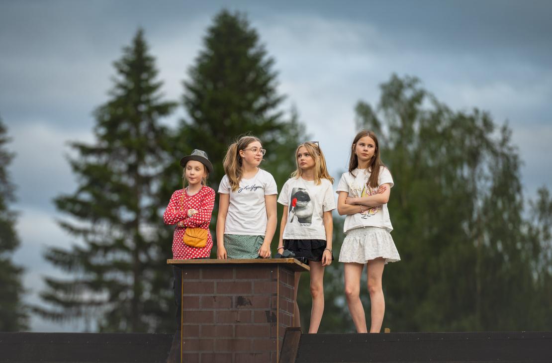 Neljä nuorta tyttöä seisoo vierekkäin tiilisavupiipun vieressä rakennuksen katolla Möhkön kesäteatterin Peitetarina-näytelmässä. Heillä on yllään kesäiset vaatteet, osa pitää käsiään puuskassa, ja he katsovat tarkkaavaisina sivuun taustallaan korkeat vihreät puut ja pilvinen taivas.