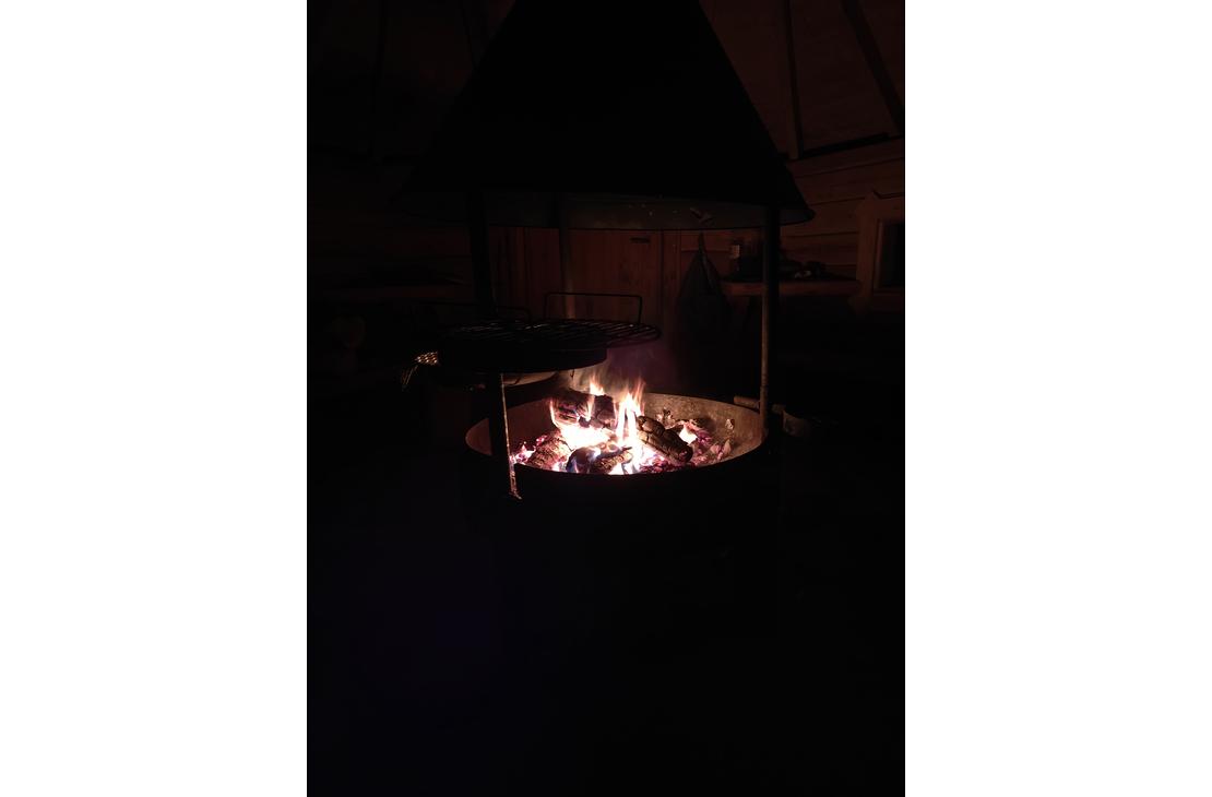 Pimeässä otettu kuva kodan tai laavun sisällä palavasta nuotiosta pyöreässä tulisijassa. Liekit valaisevat ympäröivää hämäryyttä.