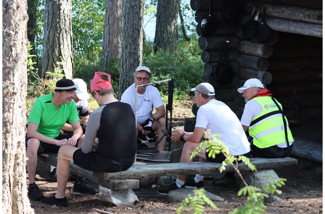 Ryhm&auml; miehi&auml; istuu puupenkeill&auml; nuotiopaikan &auml;&auml;rell&auml; mets&auml;ss&auml; sijaitsevan hirsilaavun edustalla.