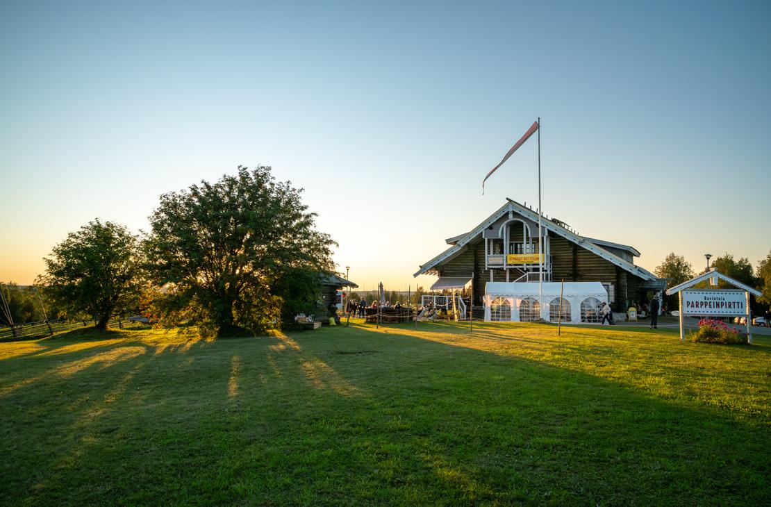 Laajakuva Parppeinpirtin suuresta hirsirakennuksesta ja sen pihapiiristä aurinkoisena alkuiltana. Aurinko luo pitkiä varjoja nurmikolle, ja ihmisiä on kokoontunut rakennuksen eteen.