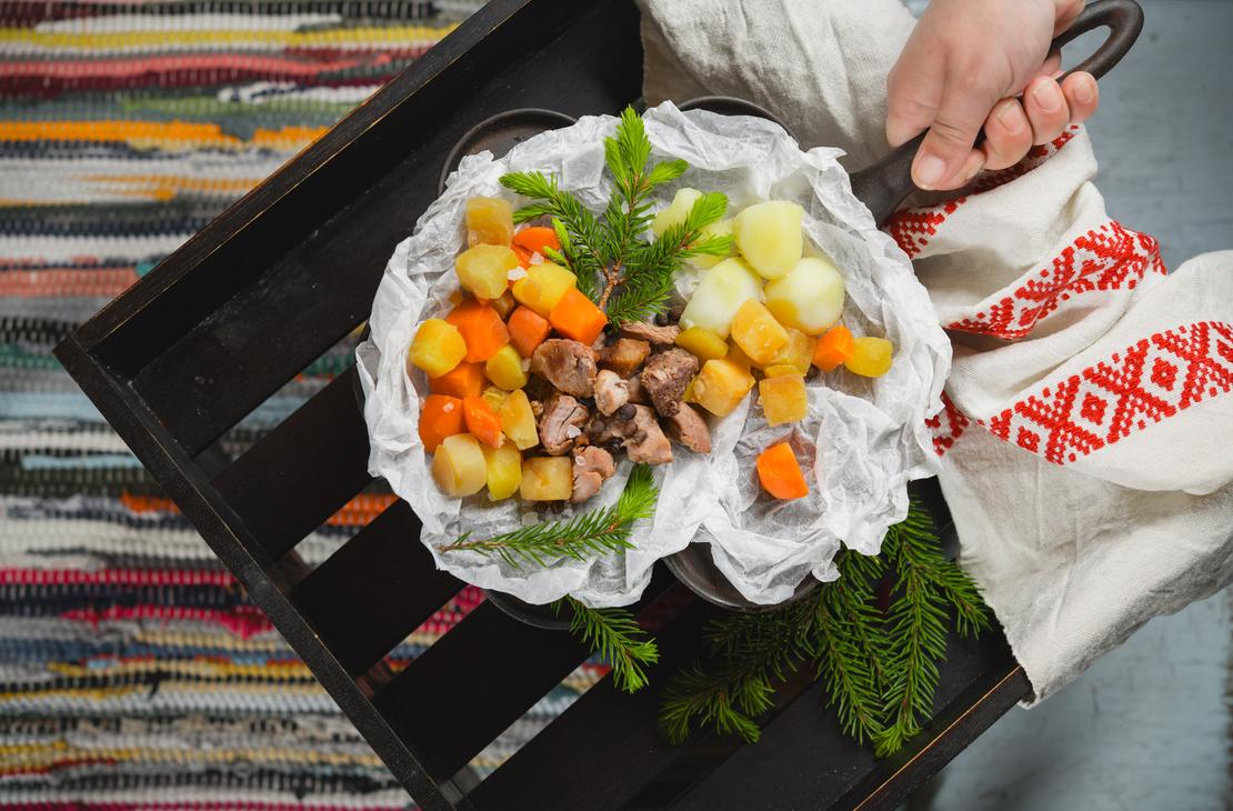 Mustassa puulaatikossa leivinpaperin päällä tarjoiltu karjalanpaisti ja keitetyt juurekset koristeltuna kuusenoksilla. Käsi pitelee valurautapannun vartta kirjotun liinan läpi Ravintola Parppeinpirtissä.