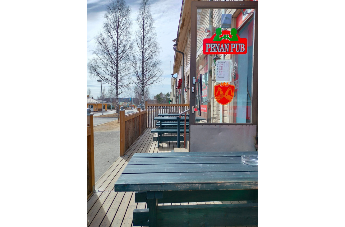 Kuva esittää Penan Pubin ulkoterassia aurinkoisena päivänä. Etualalla on puinen, tummansininen terassipöytä. Terassilla on puulattia ja tummansinisiä pöytä-penkkiyhdistelmiä. Terassin vieressä on rakennuksen lasiovi ja ikkuna, jossa on punainen, vihreä- ja kelta-oranssisävyinen "PENAN PUB" -kyltti näkyvissä. Taustalla näkyy katua, nurmikkoa ja koivuja, jotka ovat vielä lehdettömiä.