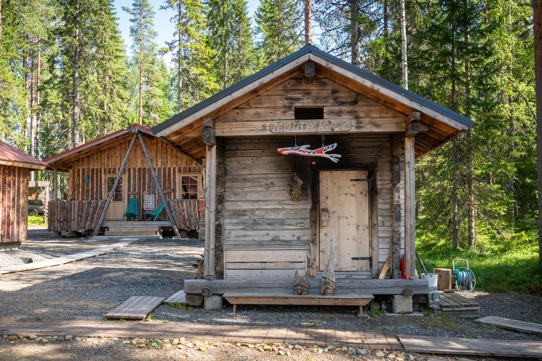 Harmaantuneesta hirrest&auml; rakennettu perinteinen sauna mets&auml;n siimeksess&auml;. Oven yl&auml;puolella on puinen kyltti, jossa lukee "MUSTA KYPY", ja terassilla on puisia koristeveistoksia.