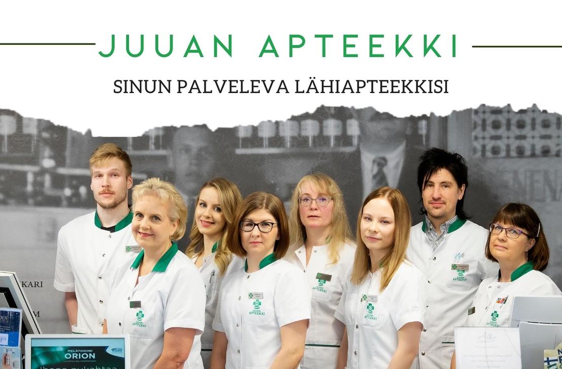 Ryhmäkuva Juuan Apteekin henkilökunnasta, jossa seitsemän naista ja kaksi miestä seisovat rivissä valkoisissa takeissa. Naiset etualalla ovat pukeutuneet valkoisiin takkeihin, joissa on vihreät kaulukset ja taskut. Taustalla on mustavalkoinen kuva tiilitalosta tai historiallisesta rakennuksesta. Kuvan yläosassa lukee vihreällä tekstillä "JUAN APTEEKKI" ja alapuolella pienemmällä tekstillä "SINUN PALVELEVA LÄHIAPTEEKKISI".