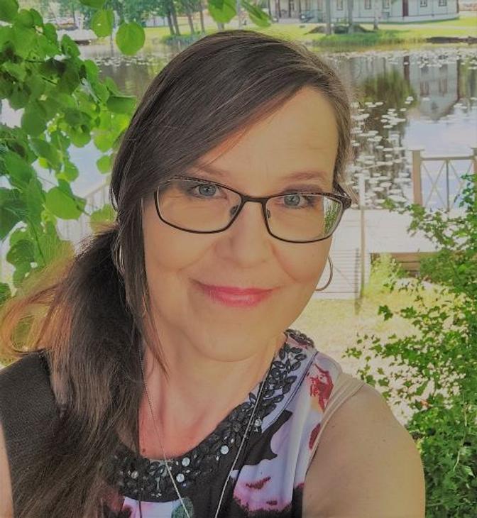 kuvassa on keski-ikäinen nainen, jolla on pitkät, ruskeat hiukset, jotka ovat osittain sidottu taakse. Hänellä on silmälasit ja hän hymyilee lempeästi kameraan. Hänellä on päällään kirjava toppi. Taustalla näkyy vihreää kasvillisuutta, mahdollisesti pensaita tai puita. Kuva on otettu ulkona ja valaistus on luonnollinen.