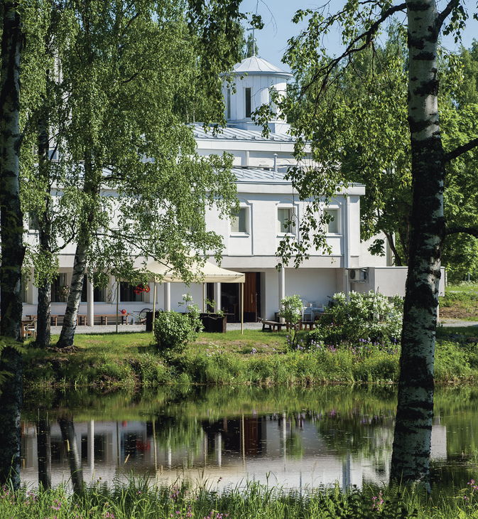 Lintulan luostarin valkoinen rakennus kuvattuna veden ääreltä kesäisenä päivänä. Etualalla on lampi, josta rakennus ja sitä kehystävät koivut heijastuvat.