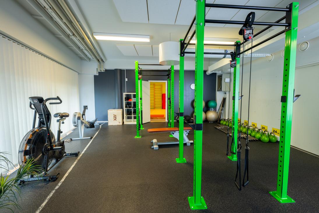 Kuva esittää Kinesiocenter Kuntoilukeskuksen harjoitustilaa. Lattiana on tumma kuminen lattia, ja tilassa on kirkkaan limevihreitä metallisia telineitä crossfit-tyyliseen harjoitteluun. Vasemmalla näkyy kuntopyörä tai ergometri. Oikealla taustalla on rivi kahvakuulia ja keskellä näkyy avoimen oven kautta seuraava huone, joka on keltainen ja punainen.