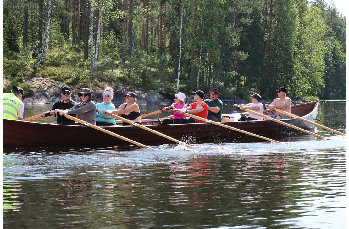 Sivukuva kirkkoveneest&auml;, jossa kahdeksan retkeilij&auml;&auml; soutaa tahdissa aurinkoisella j&auml;rvell&auml; mets&auml;isen rannan tuntumassa.