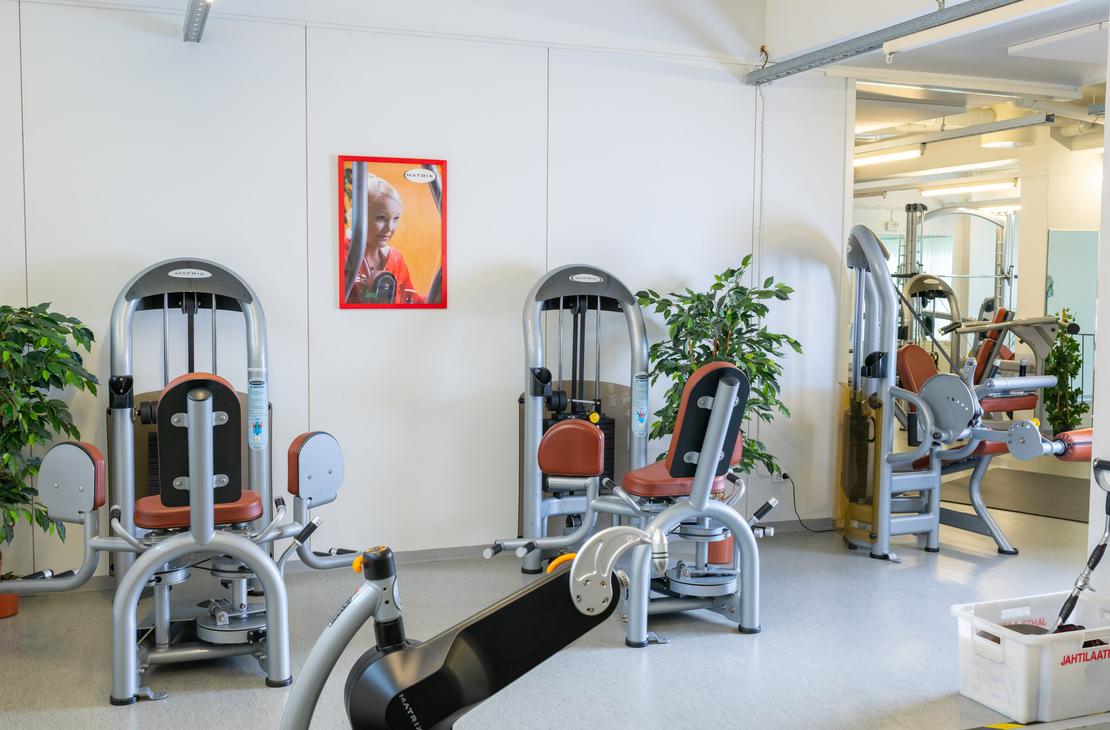 Kuva esittää osaa Kinesiocenter Kuntoilukeskuksen kuntosalitilasta, jossa on riveittäin kuntolaitteita. Laitteissa on harmaat metallirungot ja oranssinruskeat istuimet/pehmusteet. Seinät ovat valkoiset, ja niitä koristaa yksi abstrakti taulu oranssinpunaisella kehyksellä. Huoneessa on myös vihreitä huonekasveja tuomassa luonnonläheisyyttä. Lattiamateriaali on vaaleaa ja huone on hyvin valaistu.