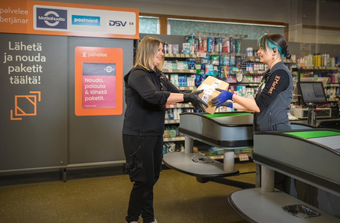 Kaksi K-Market Ilomantsin ty&ouml;ntekij&auml;&auml; ojentaa toisilleen pahvipakettia myym&auml;l&auml;n kassan ja pakettipisteen luona. Taustalla on pakettikaappi, jossa on teksti &rdquo;L&auml;het&auml; ja nouda paketit t&auml;&auml;lt&auml;!&rdquo; sek&auml; Matkahuollon, Postnordin ja DSV:n logot. Oikealla puolella n&auml;kyy kaupan kassap&ouml;yt&auml; hihnoineen ja taustalla tuotehyllyj&auml;.