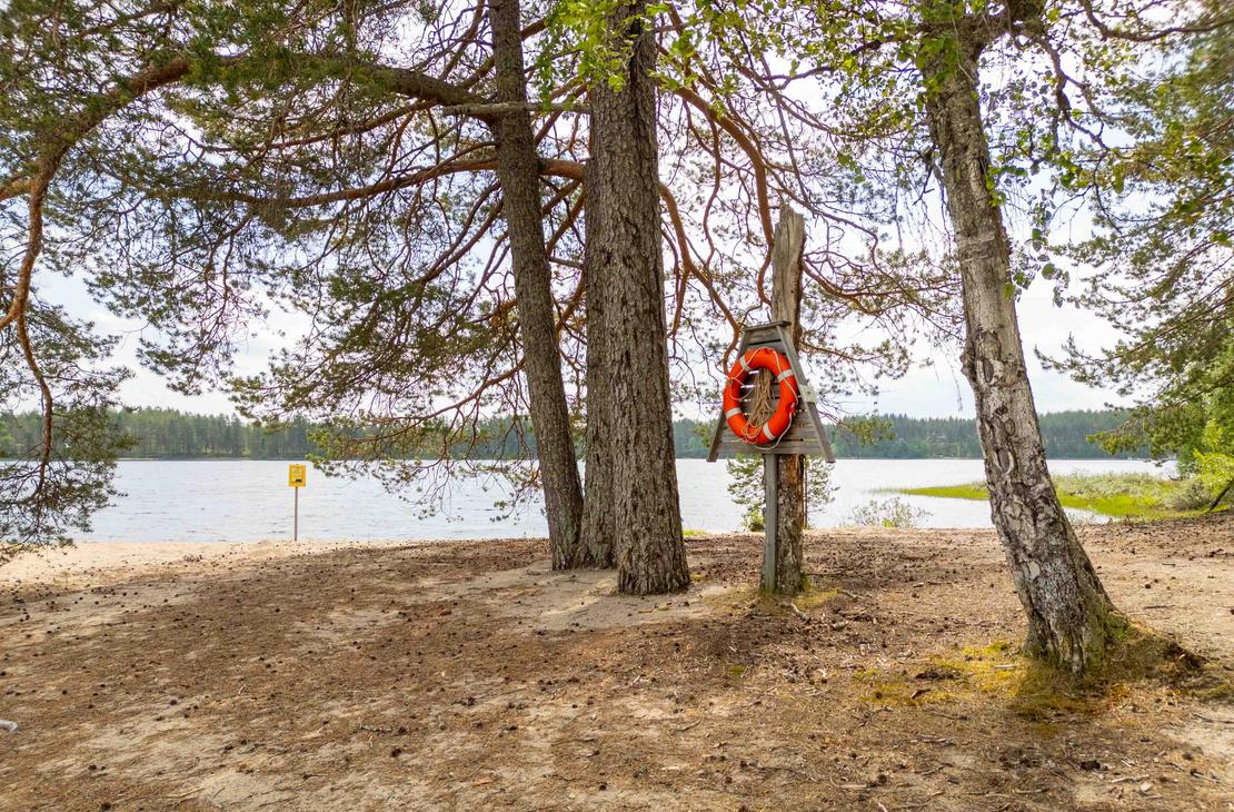 Punavalkoinen pelastusrengas kiinnitettynä puiseen telineeseen rantamännikön siimeksessä. Taustalla avautuu vaalea hiekkaranta ja tyyni järvimaisema.