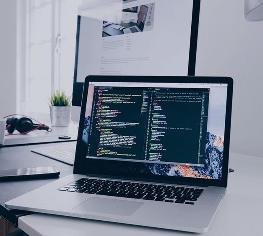 Avoin kannettava tietokone on asetettu valkoiselle ty&ouml;p&ouml;yd&auml;lle, ja sen n&auml;yt&ouml;ll&auml; on ohjelmointikoodia. Taustalla n&auml;kyy suurempi tietokoneen n&auml;ytt&ouml;, n&auml;pp&auml;imist&ouml;, kuulokkeet ja pieni viherkasvi.