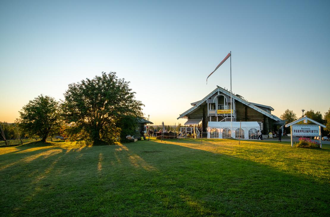 Laajakuva Parppeinpirtin suuresta hirsirakennuksesta ja sen pihapiiristä aurinkoisena alkuiltana. Aurinko luo pitkiä varjoja nurmikolle, ja ihmisiä on kokoontunut rakennuksen eteen.