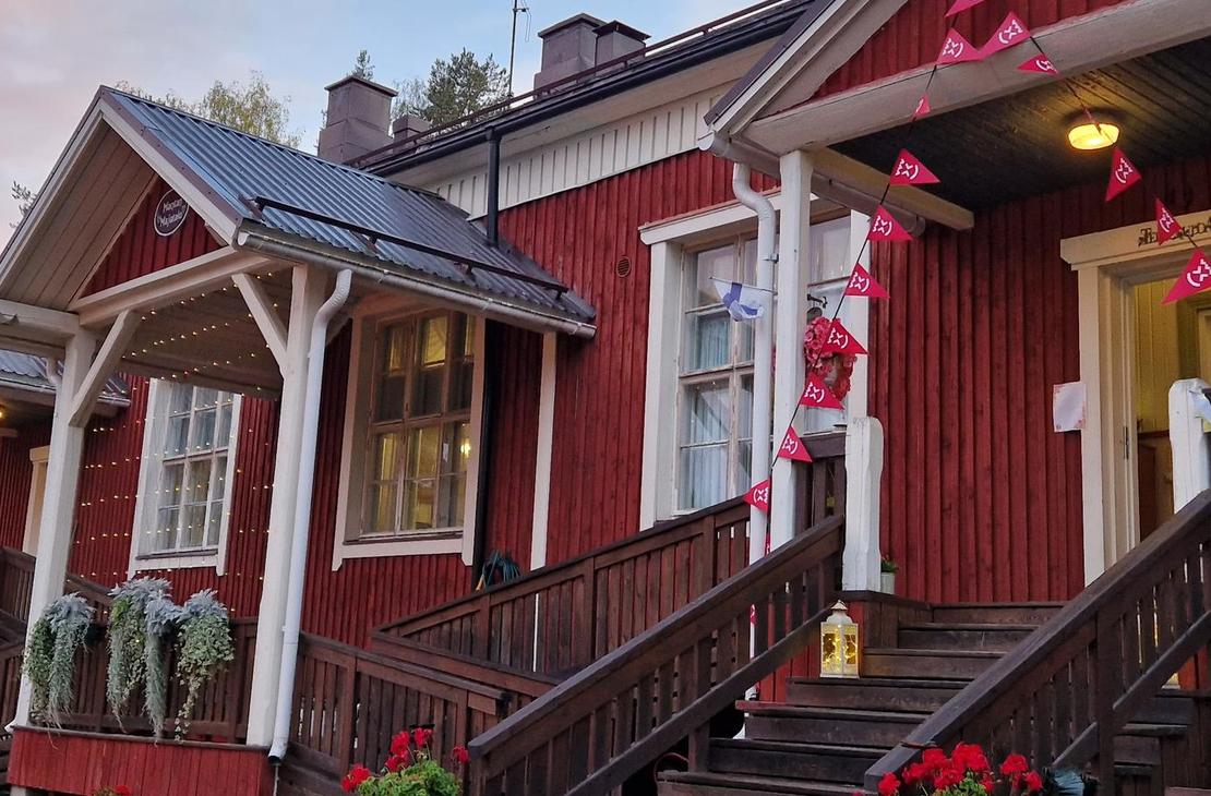 Lähikuva Mantan Majatalon sisäänkäynnistä. Punamultaisessa puutalossa on puiset portaat ja katettu kuisti, jossa on valkoiset pylväät ja kaiteet. Kuistia on koristeltu punaisilla kukilla tai nauhoilla. Sisäänkäynti on keltainen. Kuva on otettu matalalta kulmalta, ja taustalla näkyy metsän reunaa. Valo luo pitkiä varjoja, viitaten auringonlaskuun tai -nousuun.