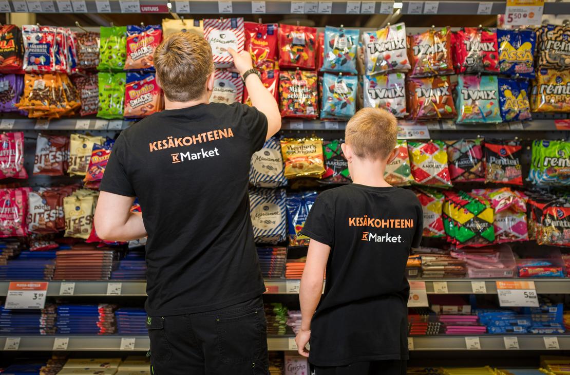 Kaksi mustiin t-paitoihin pukeutunutta henkilöä selin kuvattuna K-Market Ilomantsin karkkihyllyn edessä. Paitojen selässä on teksti ”KESÄKOHTEENA K-Market”. Vasemmanpuoleinen henkilö kurkottaa ottamaan karkkipussia ylemmältä hyllyltä, joka on täynnä värikkäitä makeispusseja, kuten TV Mix, Marianne ja Dumle.
