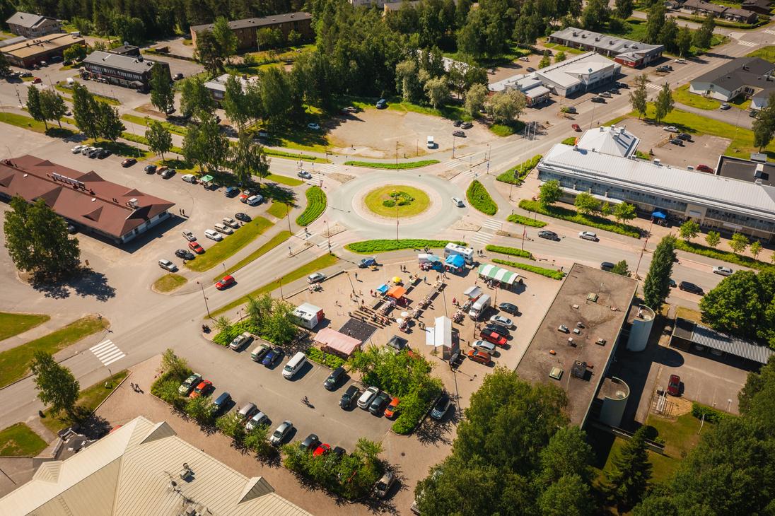 Korkealta otettu ilmakuva Ilomantsin keskustan liikenneympyrästä ja sen vieressä sijaitsevasta torialueesta, jossa on värikkäitä myyntitelttoja, ihmisiä ja ympärillä runsaasti pysäköityjä autoja aurinkoisena kesäpäivänä.