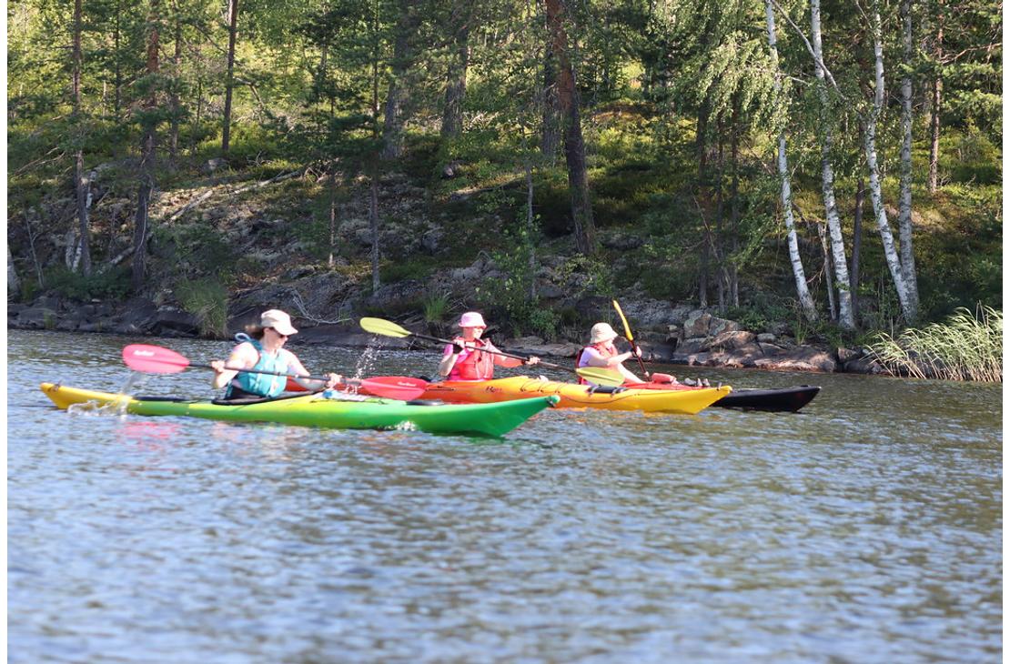 Kolme retkeilijää meloo värikkäillä kajakeilla tyynellä järvellä lähellä kallioista ja metsäistä rantaa.