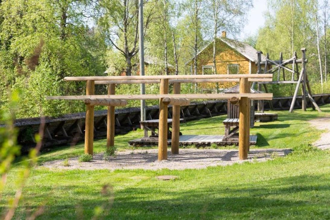 Kuva esittää Herralankosken puiston matonpesupaikan yhteydessä sijaitsevaa puista matonkuivaustelinettä. Kaksitasoinen teline seisoo hiekkapohjalla vihreällä nurmikentällä. Telineen takana näkyy vanha, keltainen puutalo ja puita, jotka ovat täydessä kesävihreydessä.