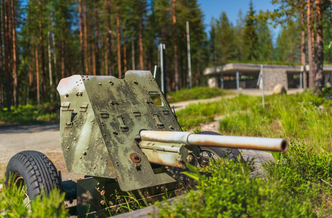 Kuva Öykkösenvaaran taistelualueen muistomerkistä tai näyttelypaikasta. Etualalla on ruosteinen, maastokuvioitu vanha tykki tai panssarintorjuntatykki. Tykki on esillä metsäisessä ympäristössä, ja taustalla näkyy epätarkkana matala, moderni, harmaasta betonista tehty rakennus, todennäköisesti taistelualueen museo tai opastuskeskus.