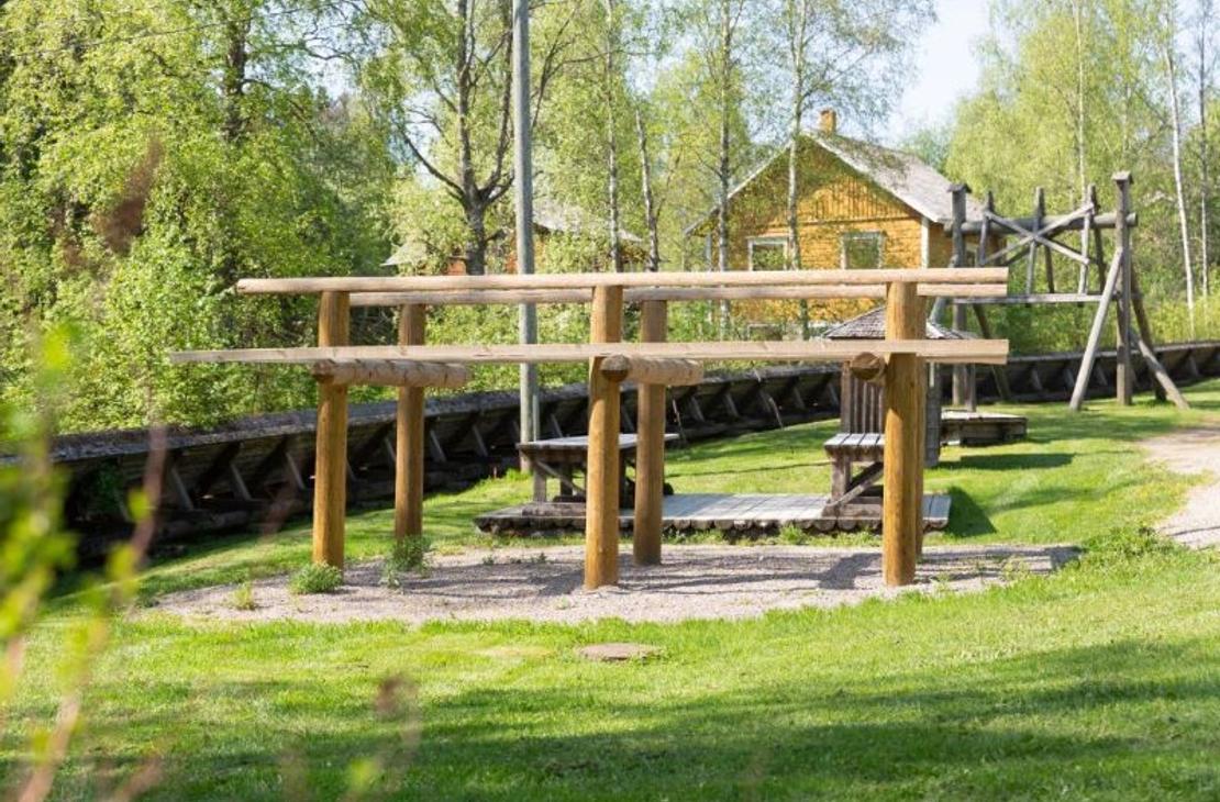 Kuva esittää Herralankosken puiston matonpesupaikan yhteydessä sijaitsevaa puista matonkuivaustelinettä. Kaksitasoinen teline seisoo hiekkapohjalla vihreällä nurmikentällä. Telineen takana näkyy vanha, keltainen puutalo ja puita, jotka ovat täydessä kesävihreydessä.