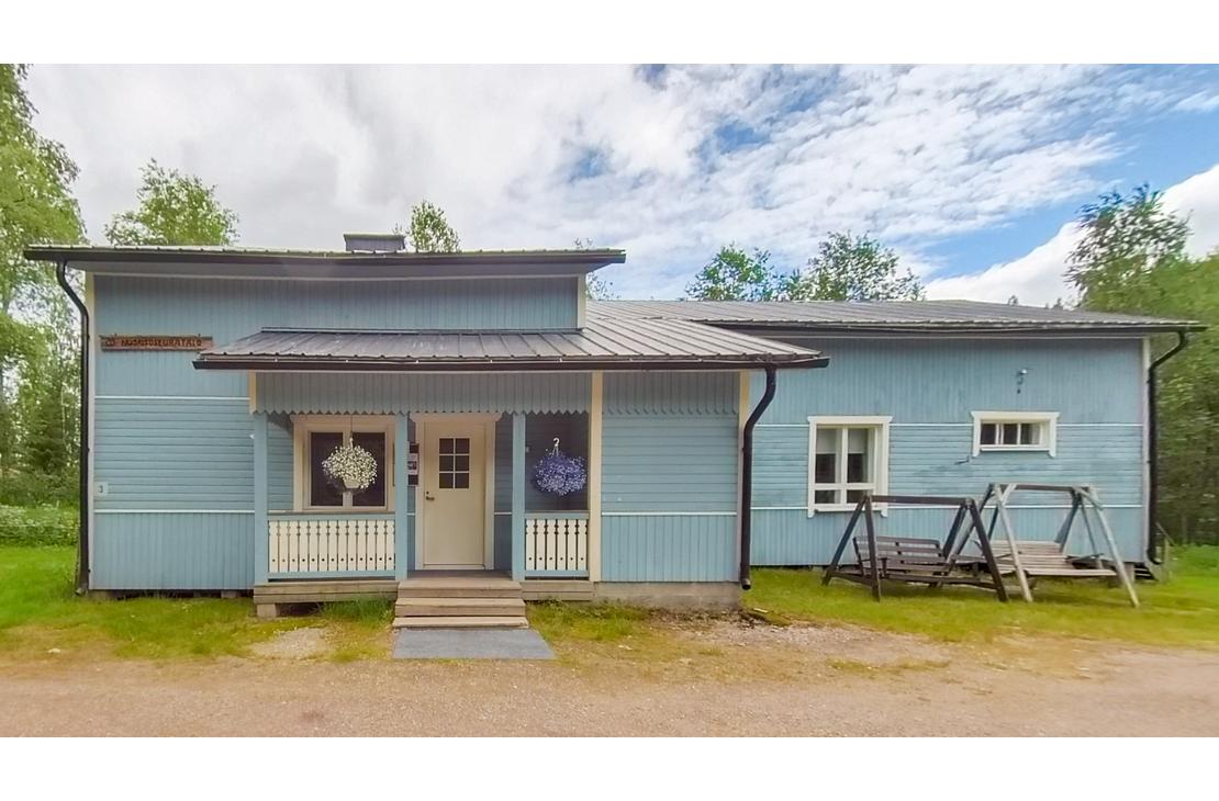 Kuva esittää matalaa, modernia, vaaleansinistä puutaloa tai rakennusta, jossa on tumma katto. Talon keskellä on pieni, katettu kuisti, jonka edessä on portaat. Kuistin oven yläpuolella näkyy pyöreä, koristeellinen valaisin tai symboli. Talon edessä on soraa tai hiekkamaata, ja oikealla sivulla on tumma, metallinen keinu tai teline nurmikolla. Taustalla näkyy rehevää kasvillisuutta ja metsää.