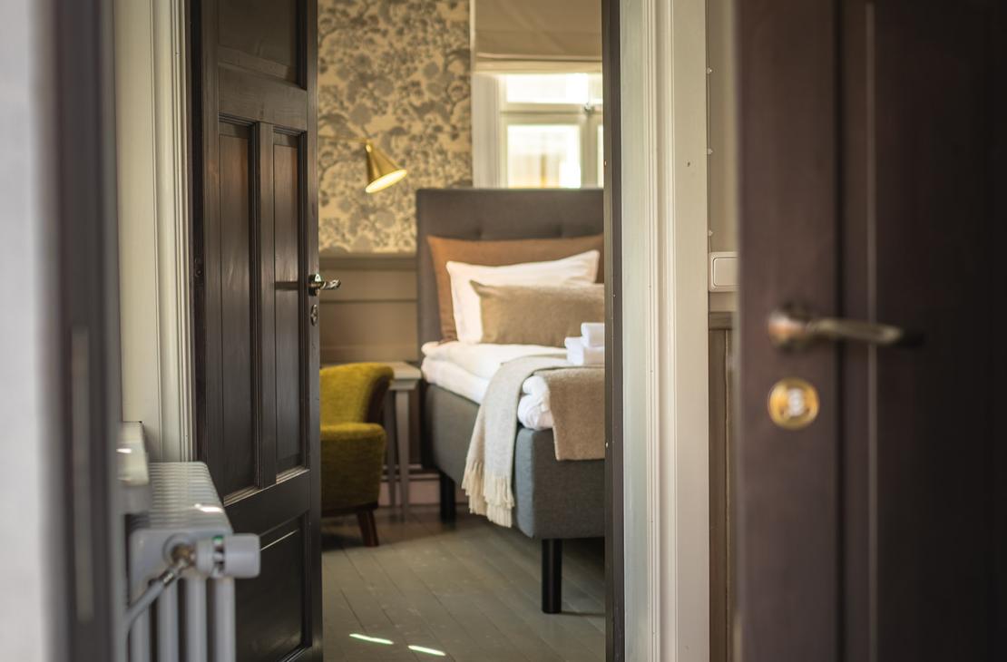 Kuva on otettu Matkailukeskus Puu-Juuka Oy:n huoneen ovelta, ja se esittää auki olevaa, tummaa puuovea, josta avautuu näkymä hotellihuoneeseen. Etualalla vasemmalla näkyy vanhanaikainen valkoinen patteri. Huoneessa, jonne ovi johtaa, on leveä sänky neutraalin sävyisillä petivaatteilla, limevihreä nojatuoli ja tumma, kuvioitu tapetti. Lattia huoneiden välillä on tummaa puuta.