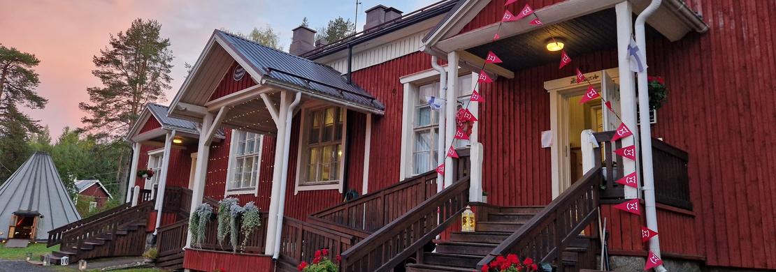 Lähikuva Mantan Majatalon sisäänkäynnistä. Punamultaisessa puutalossa on puiset portaat ja katettu kuisti, jossa on valkoiset pylväät ja kaiteet. Kuistia on koristeltu punaisilla kukilla tai nauhoilla. Sisäänkäynti on keltainen. Kuva on otettu matalalta kulmalta, ja taustalla näkyy metsän reunaa. Valo luo pitkiä varjoja, viitaten auringonlaskuun tai -nousuun.